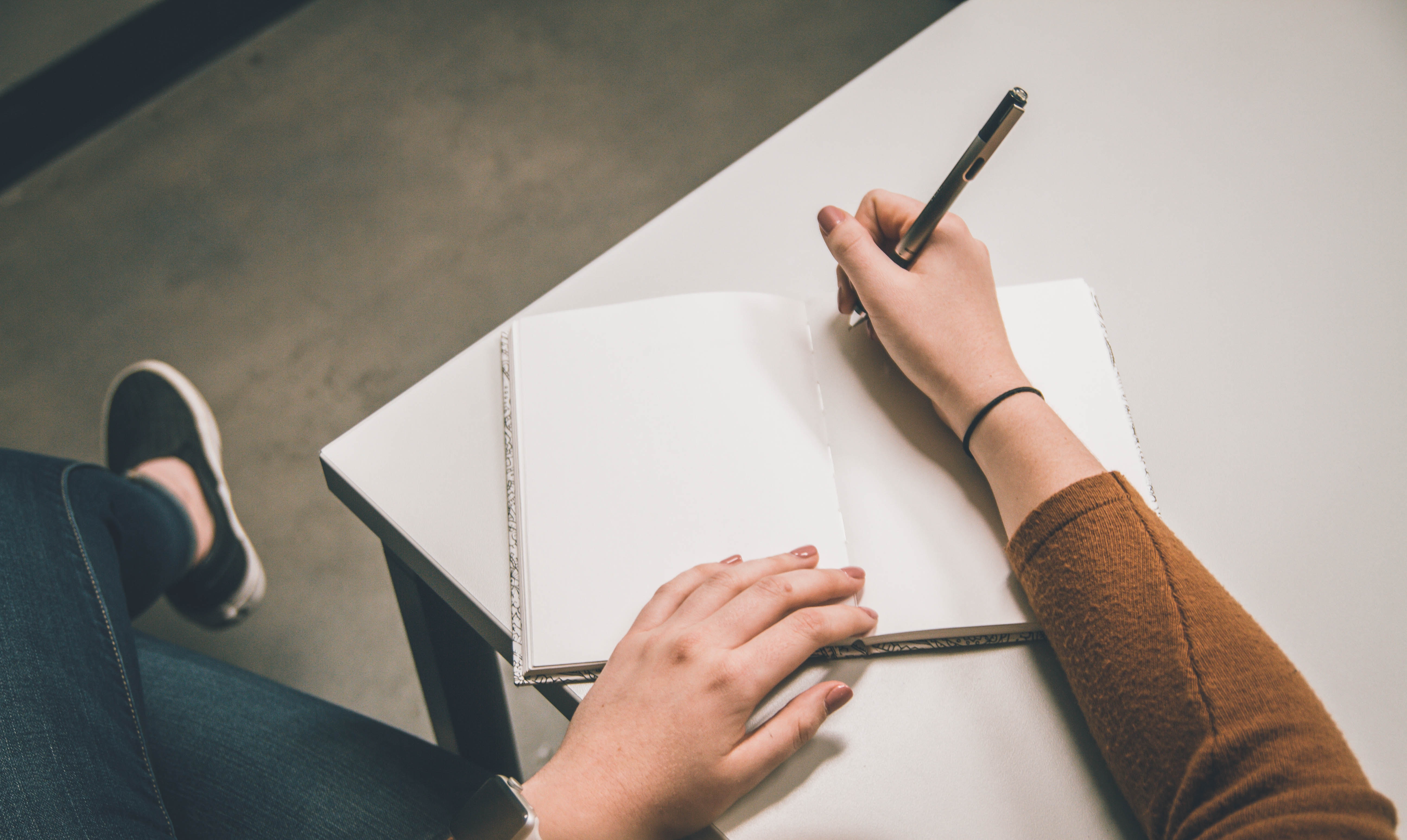 Person writing on a blank page at a table with legs crossed